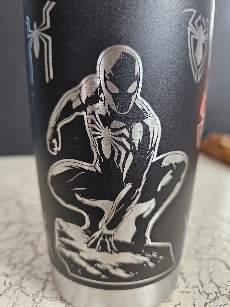 Puede incluir: Un vaso de acero inoxidable negro con una imagen grabada en plata de Spider-Man en posici&oacute;n de cuclillas. El vaso tiene una tapa plateada y un fondo plateado.