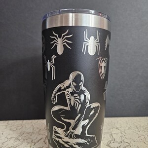 Puede incluir: Un vaso de acero inoxidable negro con un dise&ntilde;o grabado con l&aacute;ser de Spider-Man en posici&oacute;n de cuclillas. El dise&ntilde;o tambi&eacute;n incluye varios s&iacute;mbolos de telara&ntilde;a.