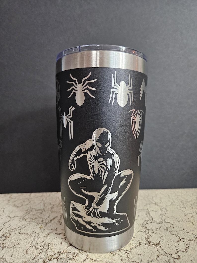 Puede incluir: Un vaso de acero inoxidable negro con un dise&ntilde;o grabado con l&aacute;ser de Spider-Man en posici&oacute;n de cuclillas. El dise&ntilde;o tambi&eacute;n incluye varios s&iacute;mbolos de telara&ntilde;a.