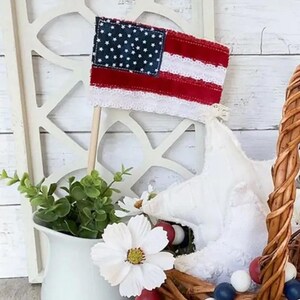 May include: A small American flag made of fabric with white lace trim is displayed on a wooden stick. The flag is red, white, and blue and has a white star pattern on the blue section. The flag is in front of a white lattice background.