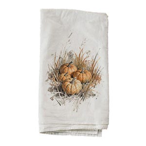 May include: A white rectangular kitchen towel featuring a central watercolor-style illustration of three orange pumpkins nestled among tall, dry brown grasses and thin stalks, rendered in a detailed, artistic, and naturalistic style.