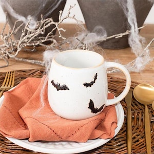 May include: A white ceramic mug with a speckled finish and three black bat silhouettes printed on the exterior. The mug features a rounded body and a curved handle, resting on a folded orange cloth napkin placed on a white plate.