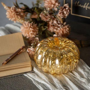 May include: A gold mercury glass pumpkin figurine with a ribbed surface and a reflective finish. The pumpkin is positioned next to an open notebook with a metallic pen resting on the pages and a bouquet of light brown flowers in the background.