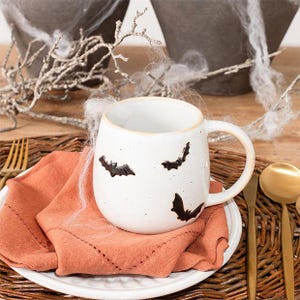 May include: A white ceramic mug with a speckled finish and three black bat silhouettes printed on the exterior. The mug features a rounded body and a curved handle, resting on a folded orange cloth napkin placed on a white plate.