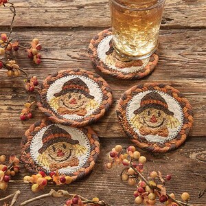 May include: Three round fabric coasters featuring embroidered scarecrow faces with brown hats and orange accents. Each coaster has a braided border in shades of brown and orange. One coaster supports a clear glass filled with a golden liquid.