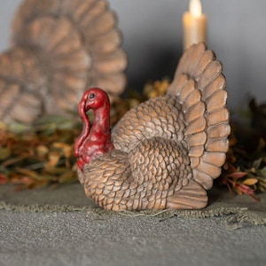 May include: A turkey figurine featuring a textured brown body with detailed feather patterns and a bright red head and neck. The sculpture is crafted with a prominent fanned tail and a realistic posture, capturing the distinct physical features of the bird.