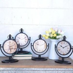 May include: Four black metal tabletop clocks with different face designs. The clocks have a round face with Roman numeral and Arabic numeral markings. The clocks are on a wooden surface with a vase of yellow and white flowers.