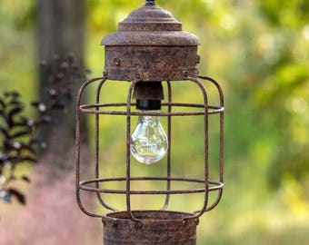 May include: A rustic, hanging outdoor lantern with a cage-like design. The lantern has a weathered, brown finish and features a clear lightbulb. The cage is made of metal bars, and the lantern is suspended by a black cord. The background is a blurred view of green foliage.