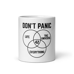 May include: White ceramic mug featuring the words "DON'T PANIC" and a Venn diagram. The diagram includes "LIFE", "THE UNIVERSE", and "EVERYTHING", with "42" in the centre. A novelty item.