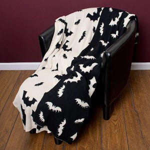 May include: A knit throw blanket featuring a two-tone design with a split color scheme of cream and black. The blanket is decorated with a repeating pattern of flying bats, where black bats appear on the cream section and cream bats on the black.