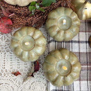 May include: Three sage green pumpkin-shaped candles with a speckled finish and a central white wick. Each candle features a rounded, ribbed design resembling a gourd. The candles are arranged on a surface with a white lace and plaid fabric.