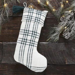 May include: A white Christmas stocking with a green and black plaid pattern. The stocking has a green tassel and a loop for hanging. The background is a dark wood surface with pine branches and lights.