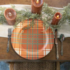 May include: A round dinner plate featuring an orange, white, and sage green plaid pattern. The plate is set on a table with a fork, a knife, and a spoon, accompanied by three brown pillar candles and a green leafy garland on a runner.