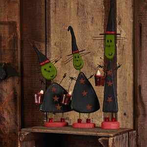 May include: Three wooden witch figures with green oval heads, black pointed hats with red glitter bands, and black triangular bodies decorated with brown stars. Each figure stands on a red base and holds small brown buckets containing candy corn.