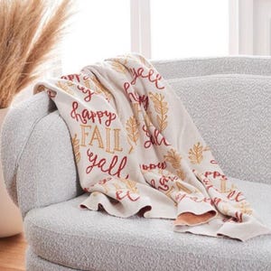 May include: A cream-colored throw blanket draped over a gray armchair. The blanket features the words "Happy Fall Y'all" in burgundy and gold lettering, along with leaf designs. The blanket's edges are folded over, revealing a light brown underside.