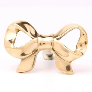 May include: A gold-toned metal bow-shaped door knob or pull. The bow is tied in a knot in the center.