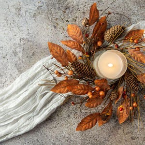 May include: A lit candle in a glass holder surrounded by an autumn-themed wreath featuring brown and orange faux leaves, pinecones, and small orange berries, all resting on a long, crinkled white fabric runner with a textured gray surface.
