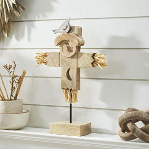May include: A wooden scarecrow figurine featuring a light brown finish, a carved face, a crescent moon cutout on the torso, and straw-like accents at the hands and base. A small silver-colored bird sits on the hat, mounted on a black metal rod.