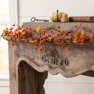 May include: A decorative garland featuring thin brown twigs, small orange berries, and yellow-orange leaves draped across a wooden mantel. The mantel is adorned with a small striped pumpkin, a metal skeleton key, and a pillar candle.