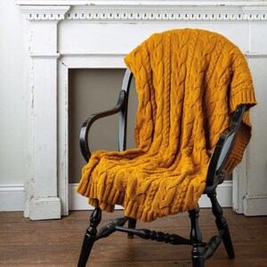 May include: A mustard yellow cable-knit throw blanket draped over a black wooden chair. The blanket's texture and color create a cozy aesthetic. The chair is positioned in front of a white fireplace.