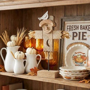 May include: A wooden scarecrow figurine with a bird on its head and a crescent moon cutout, displayed alongside two white ceramic pitchers, small white pumpkins, dried wheat stalks, amber glassware, and a sign reading FRESH BAKED PIE.