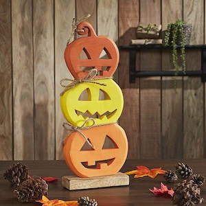 May include: A vertical stack of three carved wooden jack-o'-lanterns featuring two orange pumpkins and one yellow pumpkin in the center. Each pumpkin has cut-out facial features and is tied with rustic twine, mounted on a rectangular base.