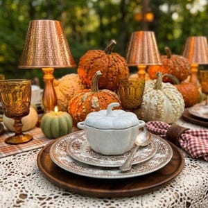 May include: A white ceramic pumpkin-shaped lidded tureen sits on a brown patterned plate and wooden charger. The table setting includes amber-colored glasses, copper-toned lamps, and various decorative pumpkins in orange, white, and green hues.