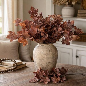 May include: A beige ceramic vase with a distressed finish holds a large arrangement of dark brown maple leaves. A separate stem of matching brown maple leaves rests on the surface in front of the vase, which is positioned near a stack of books.