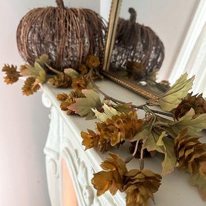 May include: A brown woven twig pumpkin decoration sits next to a garland of artificial hops and green leaves. The pumpkin has a textured, rounded shape with a small stem on top. The garland features clusters of brown hop cones and foliage.
