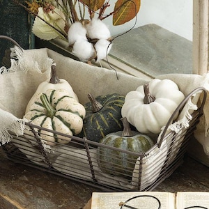 May include: A rectangular wire basket lined with a frayed beige fabric containing four decorative pumpkins in shades of white, cream with green stripes, and dark green, accompanied by a sprig of white cotton and dried leaves.