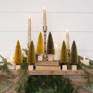 May include: A collection of six miniature bottle brush trees in shades of mustard yellow, olive green, and dark green, each mounted on a small square wooden base. The display includes three tall taper candles in gold holders and three white pillar candles.