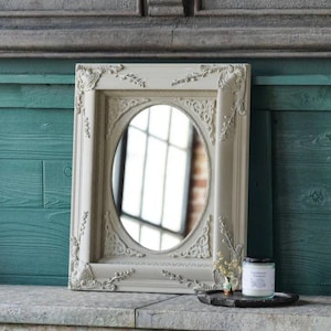 May include: A white oval mirror with an ornate, carved frame. The mirror is hanging on a wall with teal green painted wood paneling.