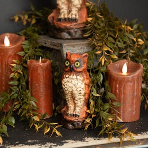 May include: A decorative owl figurine with orange and cream plumage and yellow eyes, positioned between three cylindrical brown wax candles with lit flames, surrounded by green leafy vines on a dark, weathered surface.