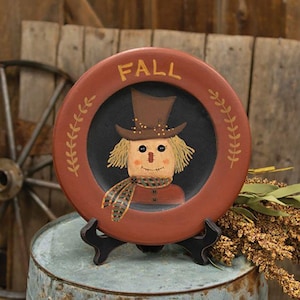 May include: A round decorative plate with a rust-colored rim featuring the word FALL in yellow lettering. The center is black and displays a painted scarecrow wearing a brown top hat and a striped scarf, accented by yellow wheat-like sprigs.