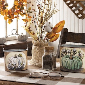 May include: Two oval metal buckets with wire handles, one featuring a white pumpkin and cotton, the other a green pumpkin with floral accents. A glass jar with a cork lid filled with dark seeds sits between them, alongside a pair of reading glasses.