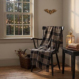 May include: A black wooden chair draped with a black and tan plaid throw blanket featuring fringe edges. The chair is positioned near a window, a woven basket filled with dried flowers, a gold lantern with a candle, and a stack of books.