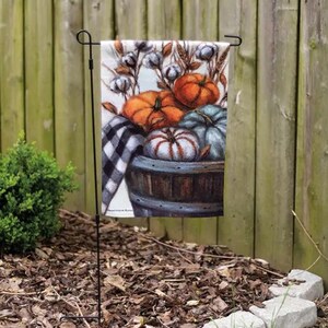 May include: A garden flag featuring a fall harvest scene. The flag displays a wooden barrel overflowing with pumpkins in shades of orange, white, and blue. Cotton stems and a black and white checkered cloth complete the autumn theme.
