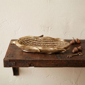 May include: A gold-colored decorative tray shaped like an ear of corn with detailed kernels and husks. Beside the corn tray are two small brown acorns and a single brass-colored skeleton key resting on the dark wooden surface.