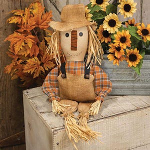 May include: A fabric scarecrow doll featuring a tan burlap hat, a tan burlap face with black button eyes, a stitched mouth, and a brown fabric nose. The doll wears an orange and blue plaid shirt with tan burlap overalls and straw accents.