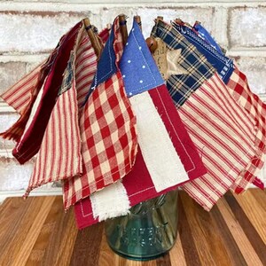 May include: A collection of small fabric flags in red, white, and blue, with various patterns, including gingham, stripes, and stars. The flags are tied together with twine and arranged in a bouquet.