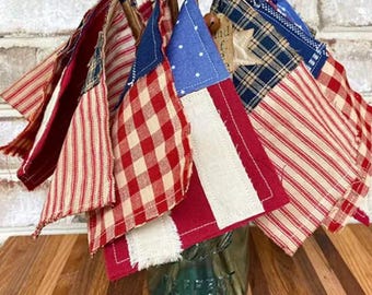 May include: A collection of small fabric flags in red, white, and blue, with various patterns, including gingham, stripes, and stars. The flags are tied together with twine and arranged in a bouquet.