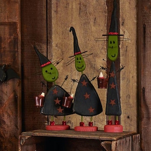 May include: Three decorative witch figures crafted from wood, each featuring a green oval face, a tall black pointed hat with red glitter trim, and a black dress adorned with brown stars. Each figure stands on a red base and holds small buckets.