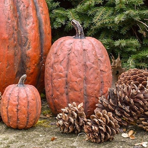 May include: Three orange pumpkins with a textured, weathered surface and dark stems, arranged alongside two large brown pinecones. The pumpkins vary in size, with one large, one medium, and one small, all featuring deep vertical ridges.