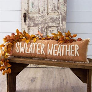 May include: A rectangular tan lumbar pillow featuring the white embroidered text "SWEATER WEATHER" in a textured, raised font. The pillow has frayed, decorative edges and is adorned with a garland of artificial autumn leaves in orange and brown.