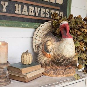 May include: A decorative turkey figurine with a red head and textured brown and cream feathers, positioned next to a stack of two vintage books topped with a small beige pumpkin, a pillar candle in a wooden holder, and dried hops.