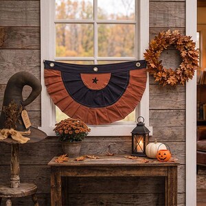 May include: A semi-circular pleated fabric bunting featuring a black top band with three brass grommets and a rust-orange lower section with a central black star. The bunting is displayed across a window frame with a rustic wooden wall background.