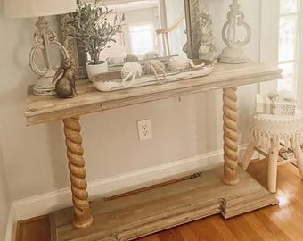 May include: A light wood console table with a whitewashed finish and rope-style legs. The table is decorated with a white ceramic lamp, a white ceramic bunny figurine, a potted plant, and a white tray with white yarn balls. The table is set against a white wall with a white baseboard.