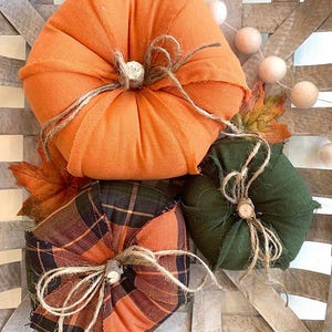 May include: Three fabric pumpkins in a woven basket. One is solid orange, one is solid dark green, and one features an orange and black plaid pattern. Each pumpkin is topped with a small wooden stem and tied with natural jute twine string.