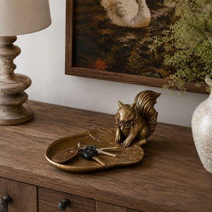 May include: A gold-toned metal trinket tray shaped like a leaf, featuring a sculpted squirrel figure at one end. The tray holds a set of metal keys with a brown leather keychain tag, providing a functional storage space for small personal items.