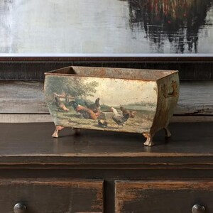 May include: A rustic metal planter with a distressed finish features a vintage illustration of chickens in a farmyard setting. The planter has four short legs and a rectangular shape.